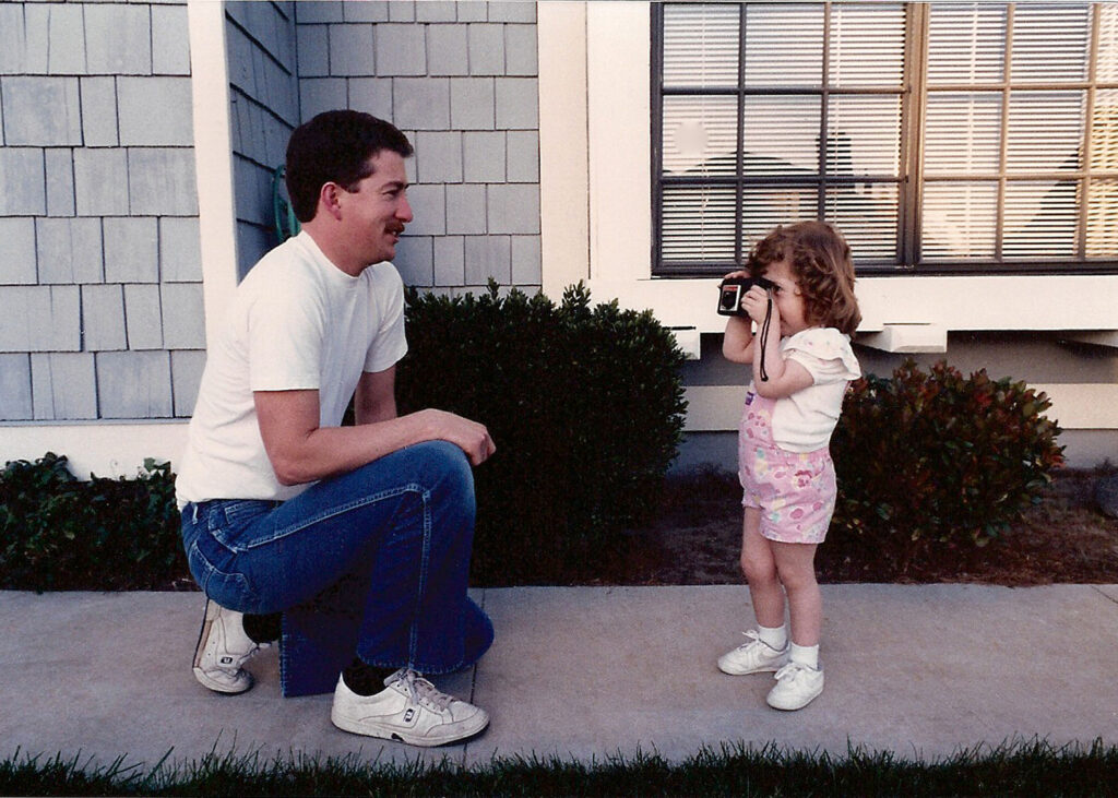 photo of young girl taking a picture of her kneeling dad with her camera in front of a house A young Jaclyn, about three years old, stands on a sidewalk holding a toy camera "photographing" her dad, who kneels in front of her smiling. They’re outside a home with gray shingles and a big window—early signs of a photographer in the making.