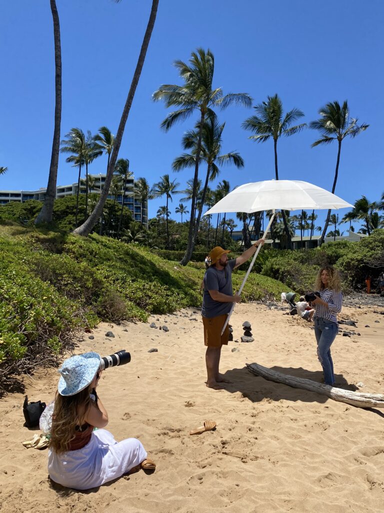 Jaclyn photographing a branding client on a tropical beach in Hawaii, assisted by her team holding a sun umbrella, showcasing her behind-the-scenes creative process.