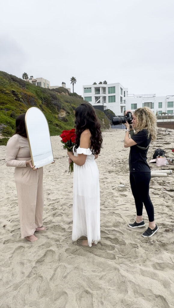 Jaclyn photographing a creative, reflective branding photoshoot on a beach, directing a client holding roses with an assistant positioning a mirror.