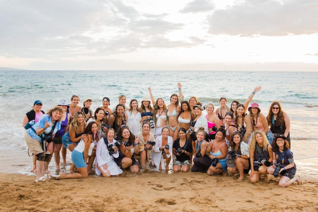 Group of smiling women photographers, including Jaclyn, posing joyfully on a sandy beach during a photography retreat in Hawaii, celebrating community and creativity.