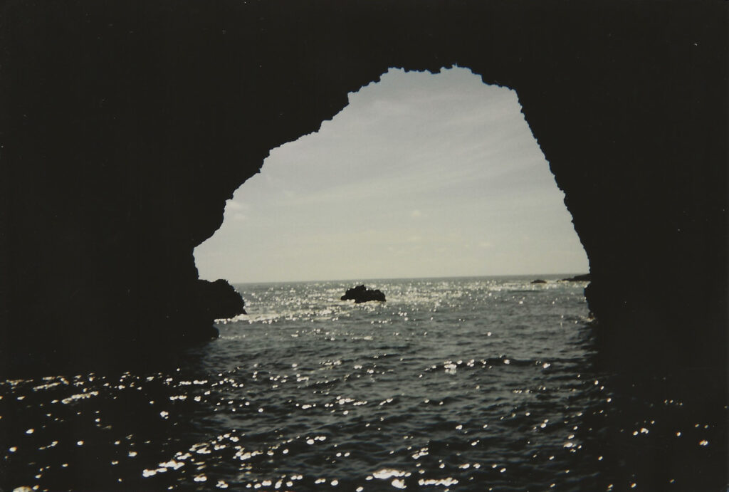 View of the ocean sparkling in the sunlight, framed through the dark silhouette of a natural rock arch on Anacapa Island. The photo, shot on 35mm film, captures the peaceful, timeless beauty of the sea.