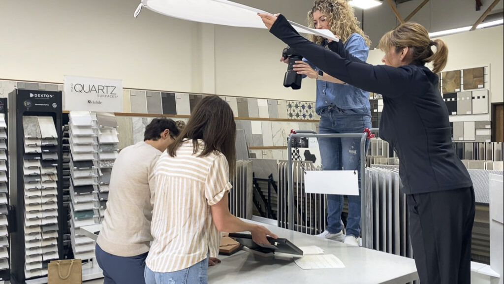 Jaclyn standing above a table capturing detail-oriented product branding images for a home design team in a tile store, assisted by her creative team to ensure perfect lighting.