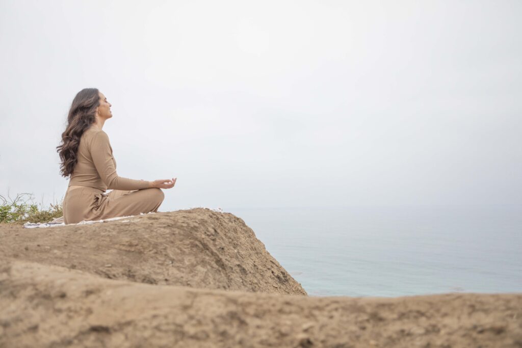 Kristine King meditating cross-legged on a cliff, facing the Pacific Ocean in peaceful stillness.