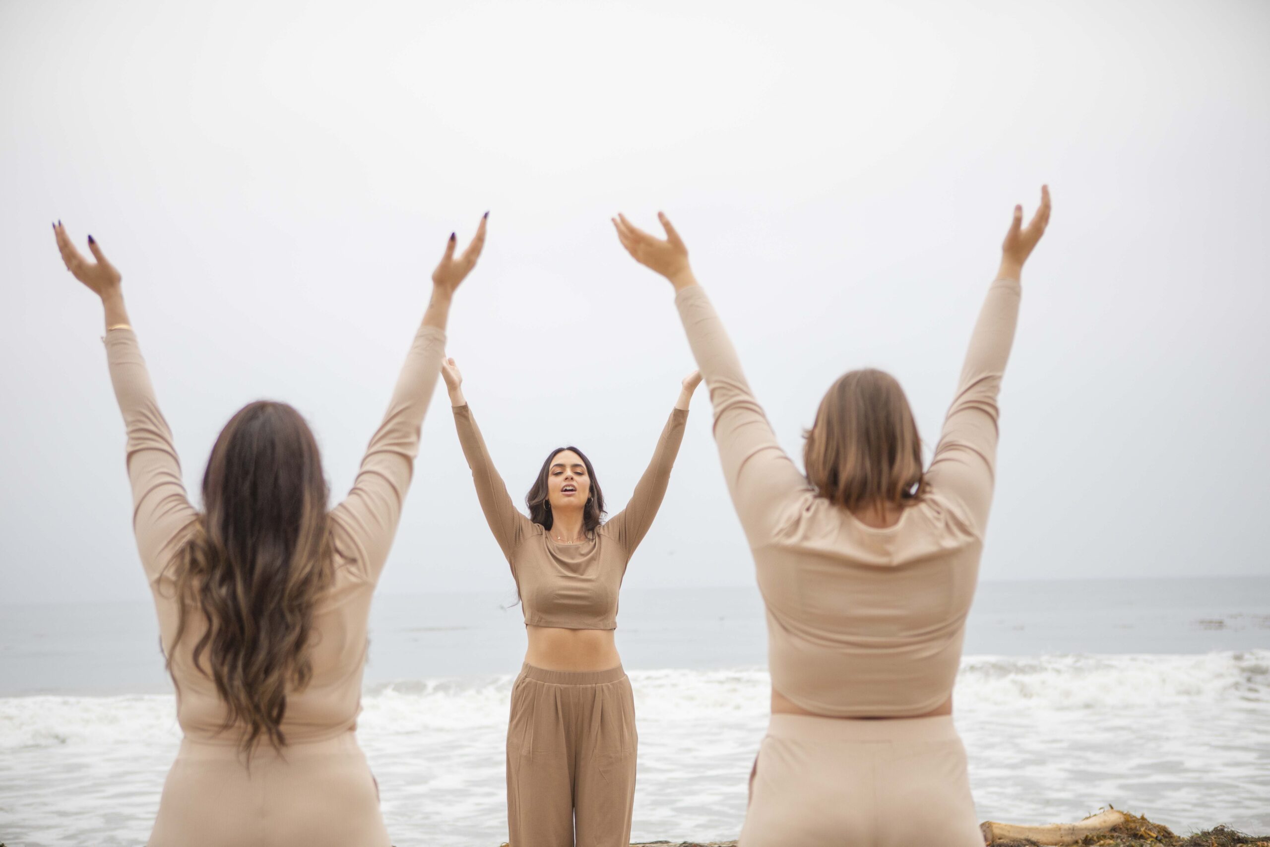 Kristine King leads two women in a beachside empowerment ritual with arms raised toward the sky.