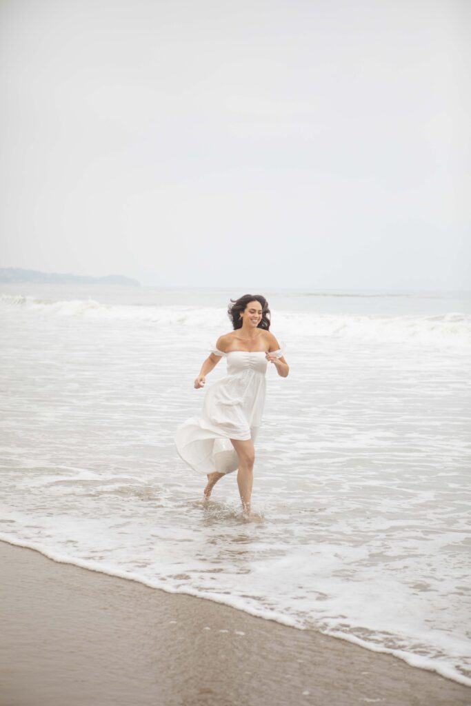 Kristine King runs barefoot through the ocean surf, smiling freely in a white dress.