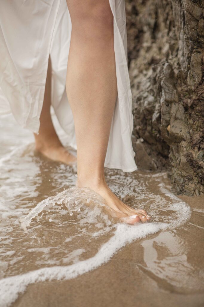 A close-up of Kristine’s bare feet stepping into the ocean water at the base of a rock.
