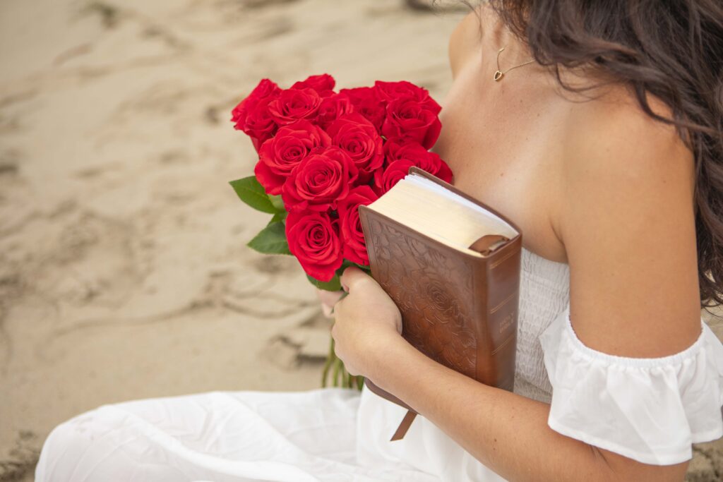 Kristine sits on the beach holding a bouquet of red roses and a leather-bound Bible against her heart.