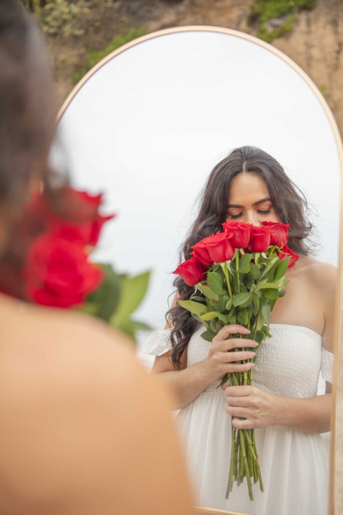 Kristine King, seen through a mirror, gently smells red roses with eyes closed in reflection.